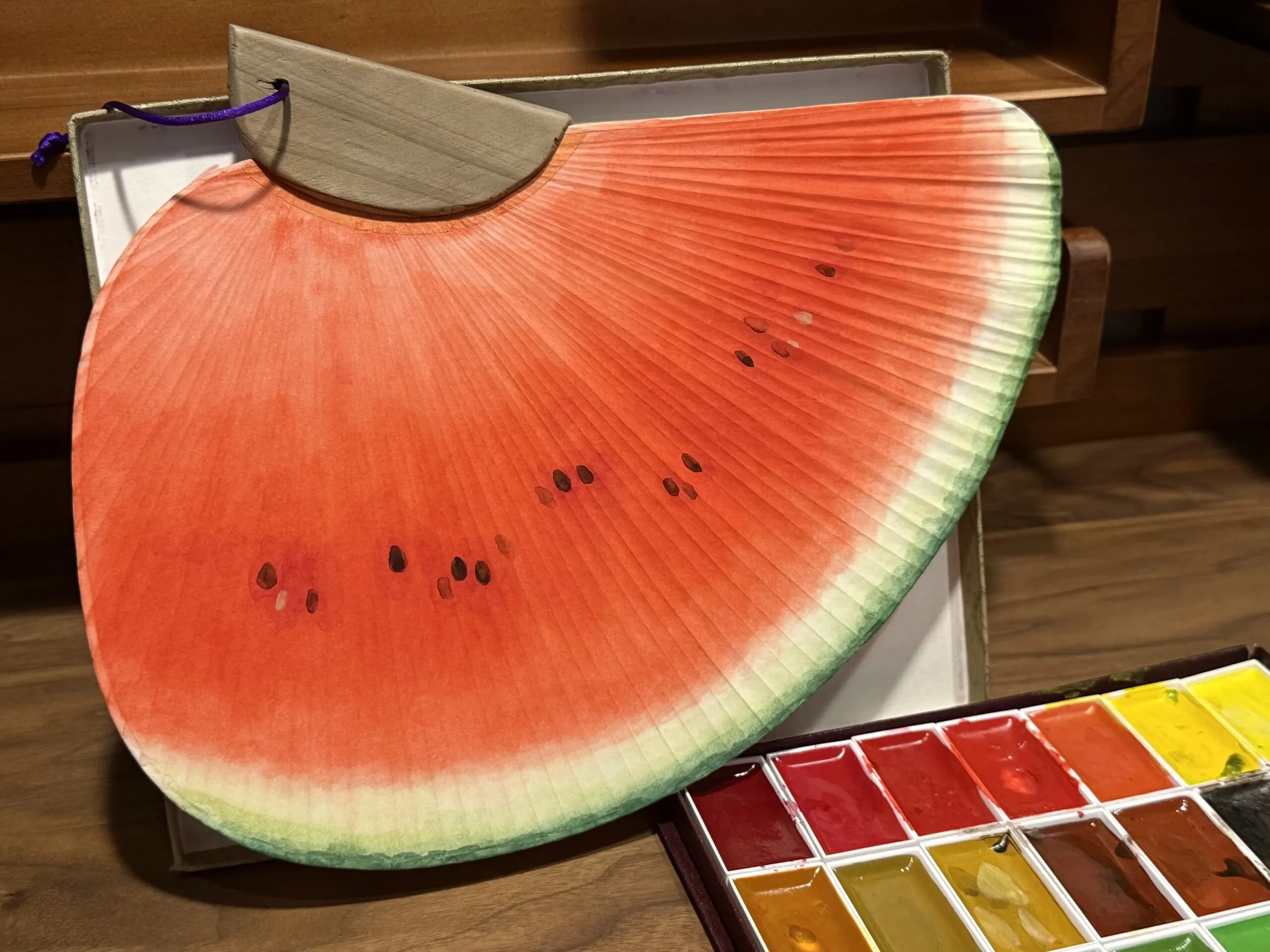 スイカの絵が描かれた千鳥うちわと、傍らに置かれた顔彩のパレット